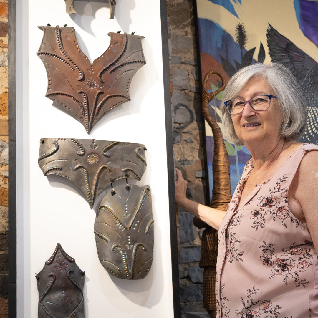 Brenda Sutton Mader, with short gray hair and glasses stands next to a white display board showcasing four medieval-style metal armor plates or breastplates in bronze and brown tones. made from clay.