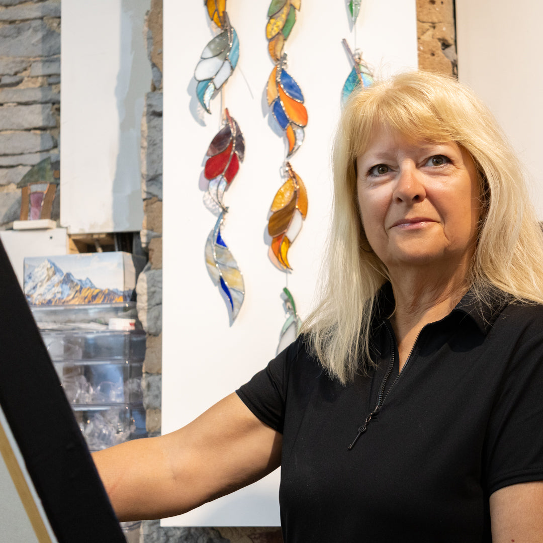 Artist Carole Renaud, wearing  a black polo shirt posing with one of her artworks in the PMAC Studio with a collection of colorful hanging stained-glass feather decorations. She is  looking at artwork. The historic grist mill's  stone-textured wall  is visible in the background.