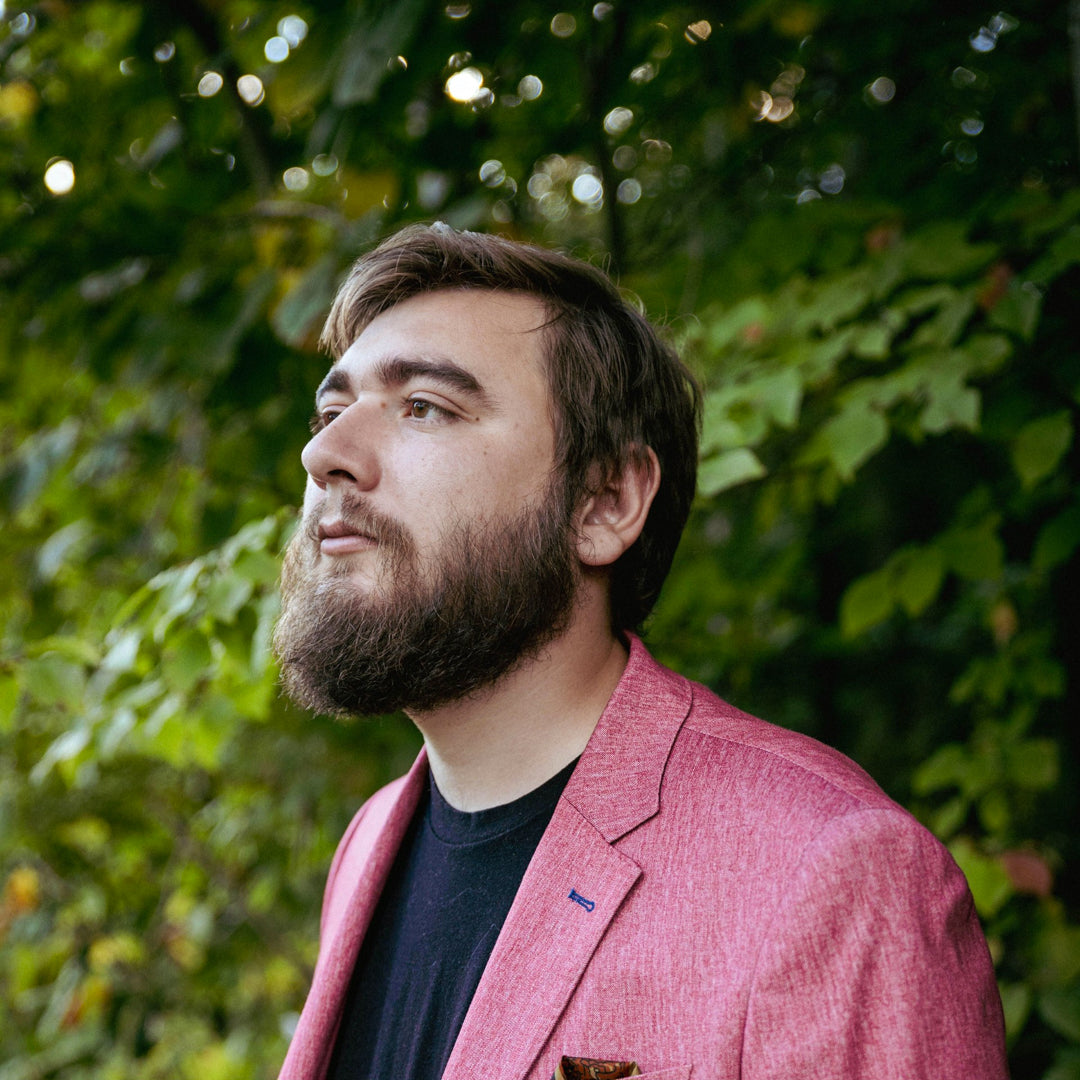 Artist Michael Poitras sporting a beard wearing a textured pink blazer over a black t-shirt, photographed outdoors against a background of green foliage.