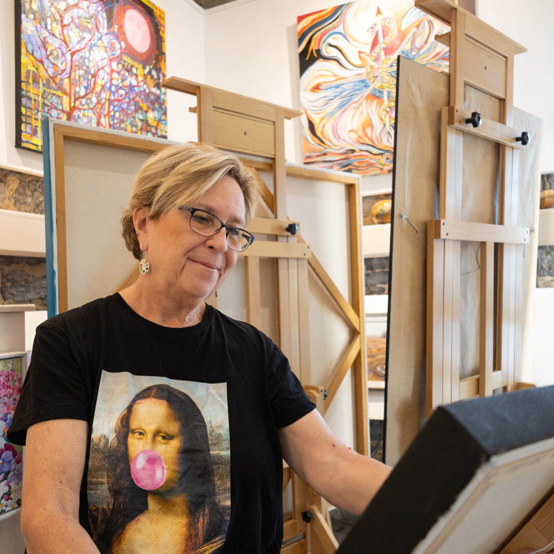 Olga Harhaj with short blonde hair and glasses, wearing a black t-shirt with a graphic print of the Mona Lisa blowing a pink bubblegum bubble, stands in the PMAC Art Gallery surrounded by easels and colorful paintings.
