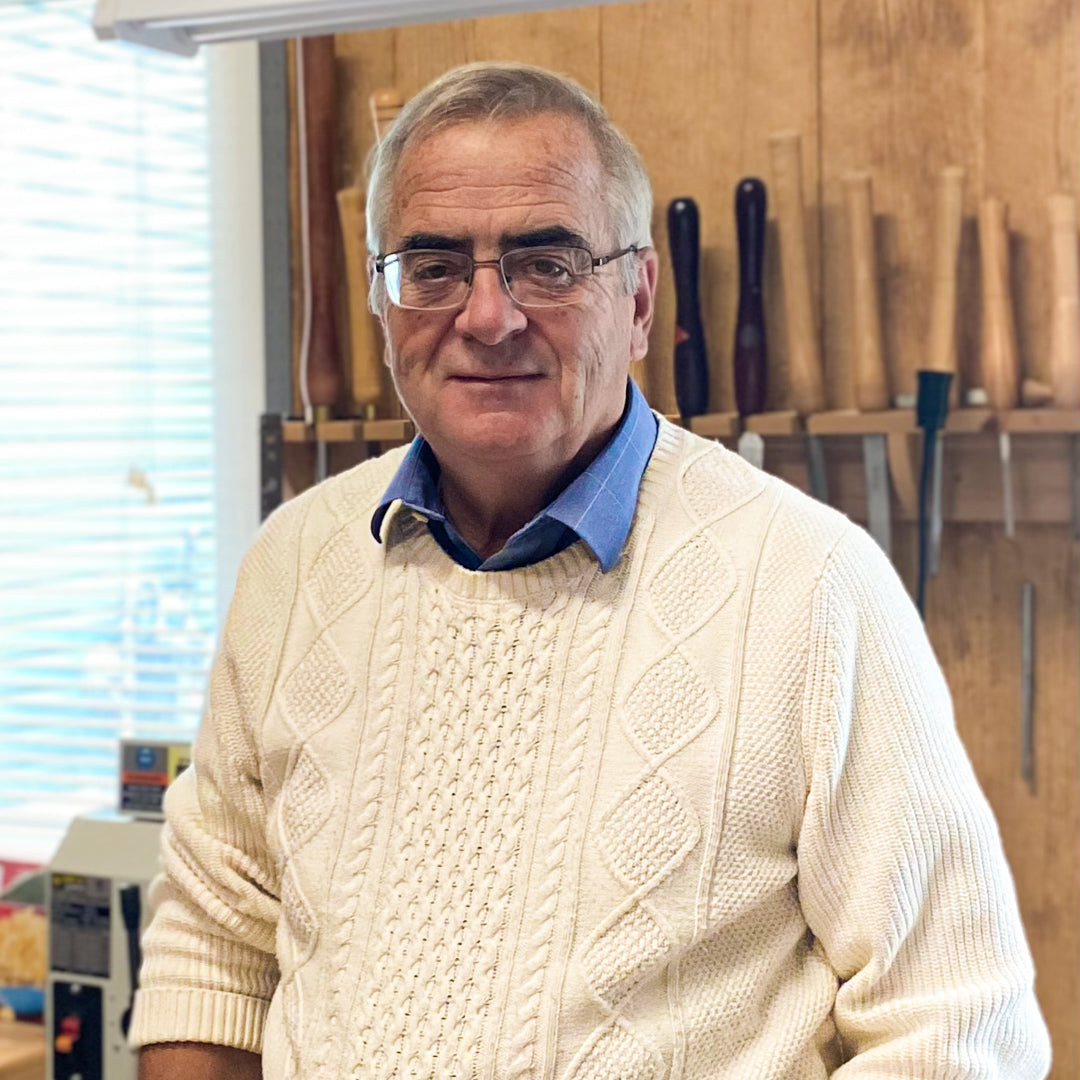Wood turner and artist, Roger Mattiuz,  wearing a cream cable-knit sweater over a blue collared shirt, standing in his woodworking shop with chisels and hand tools visible in the background.