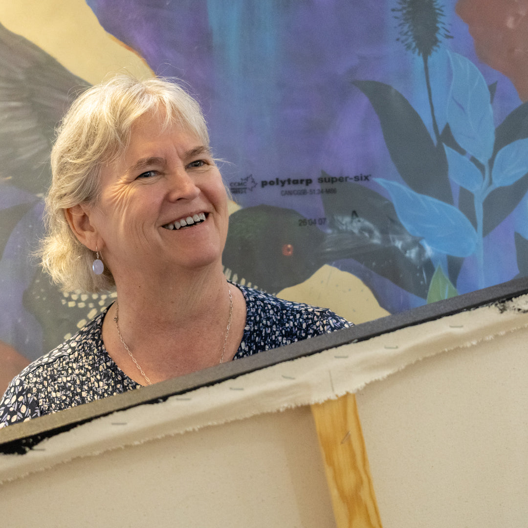 A smiling Victoria Alexander with short blonde hair is shown from the chest up, wearing a dark patterned top. She is positioned in front of one of her oversized  colorful paintings featuring a loon and blue leaves, with a large off-white canvas or board partially obscuring the foreground.